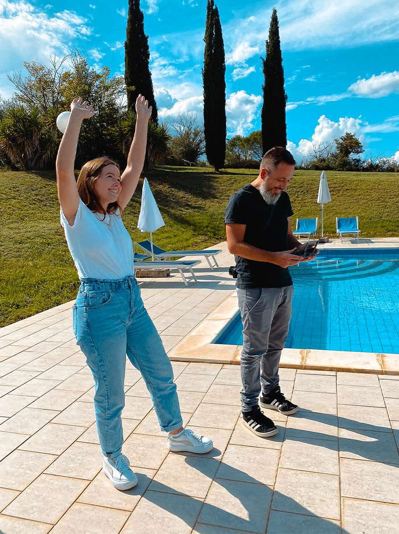 Arne und Esther stehen am Pool in Italien. Arne steuert eine Drohne während Esther die Arme hochreißt.