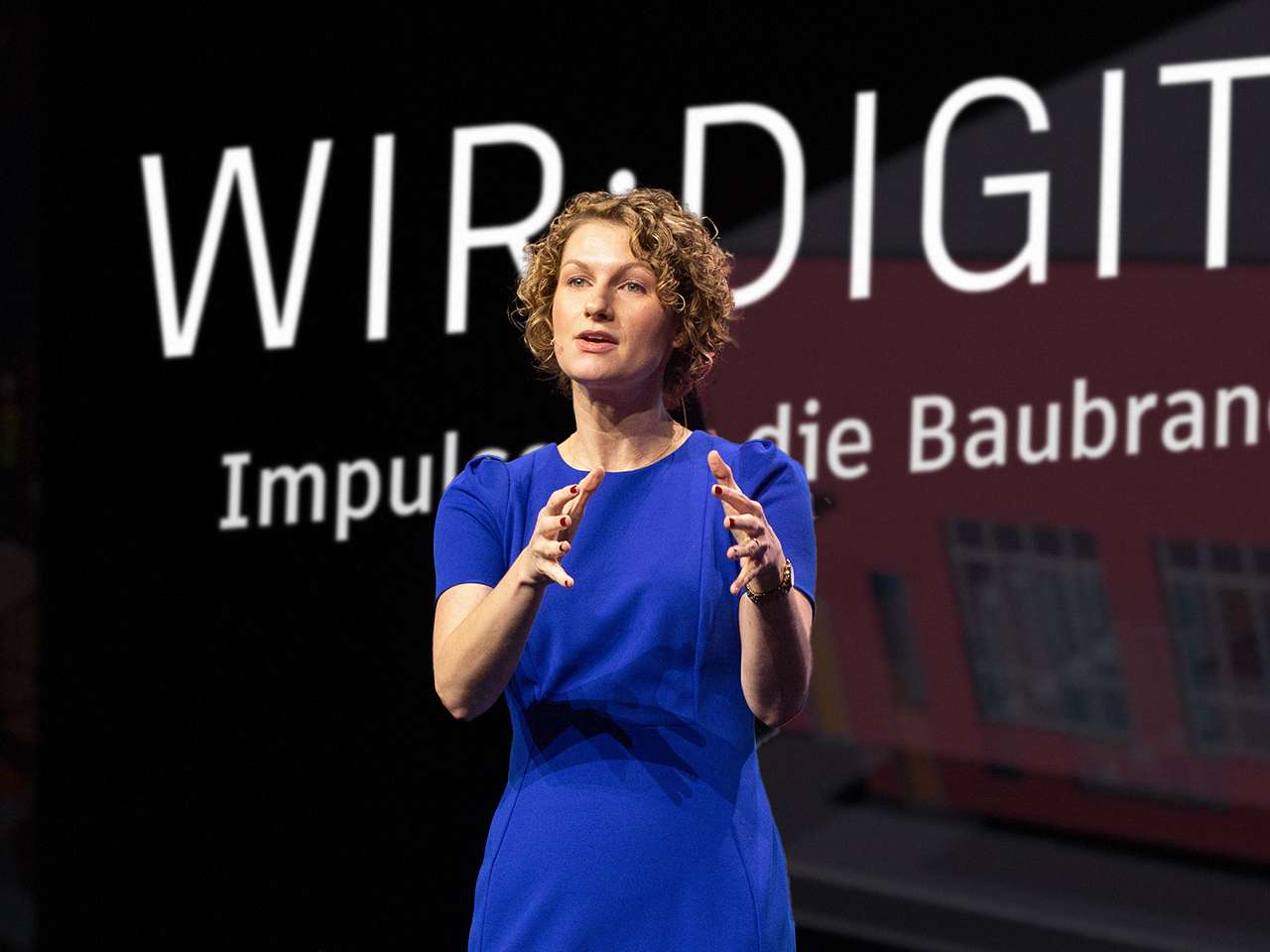 Eine Frau im blauen Kleid hält auf einer Messe einen Vortrag zu Impulsen für die Baubranche und gestikuliert mit den Händen.