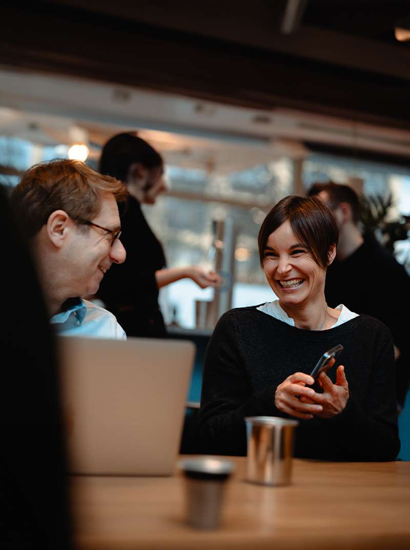 Caro und Lothar sitzen gemeinsam im Coworking-Space und unterhalten sich. Lothar hat ein MacBook vor sich stehen, Caro hält ein Handy in der Hand. Beide lachen.