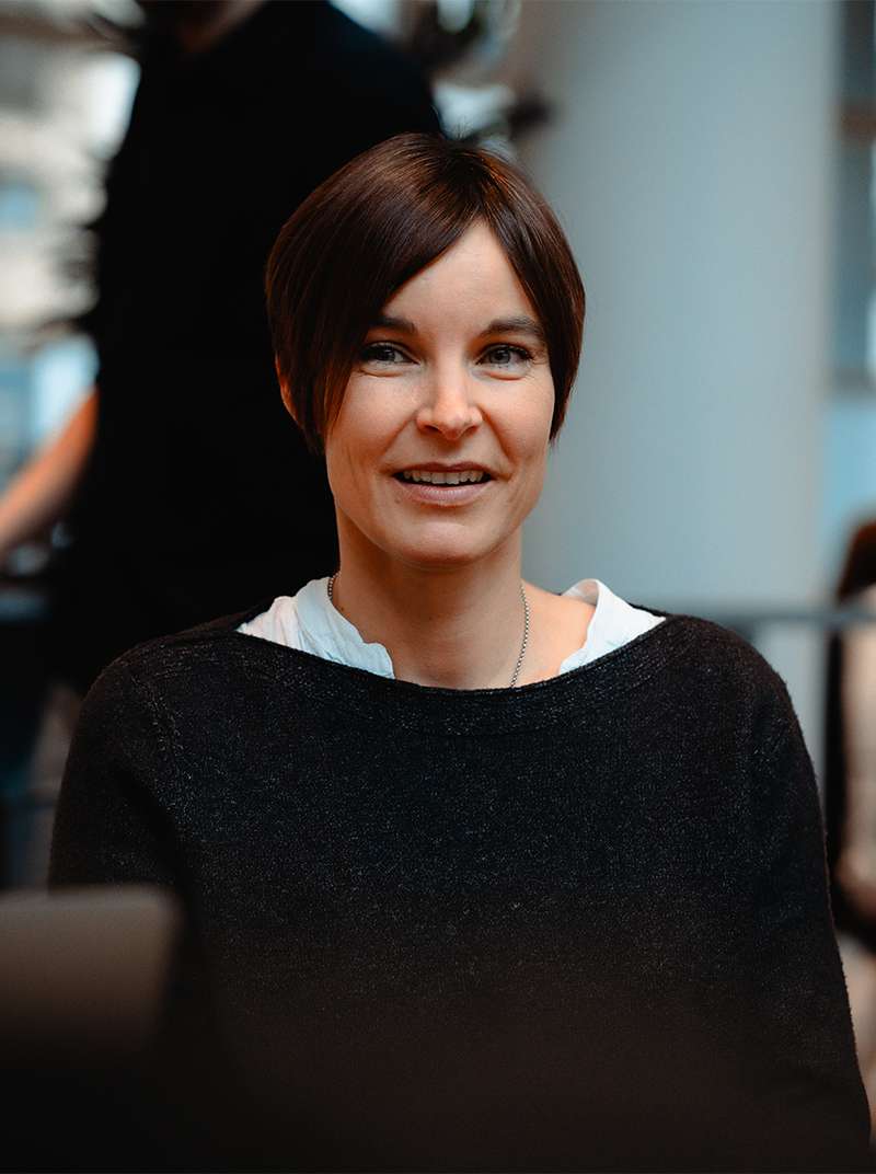 Caro schaut lächelnd in die Kamera. Sie trägt einen schwarzen Pullover und ein kragenloses weißes Hemd darunter.