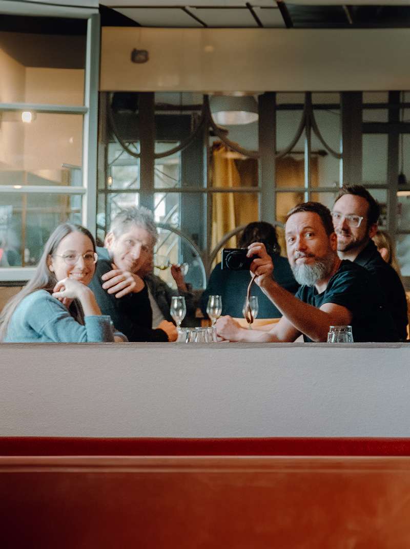 Friedi, Arne, Sarah und Felix sitzen gemeinsam an einem Restauranttisch und schauen lachend in die Kamera.