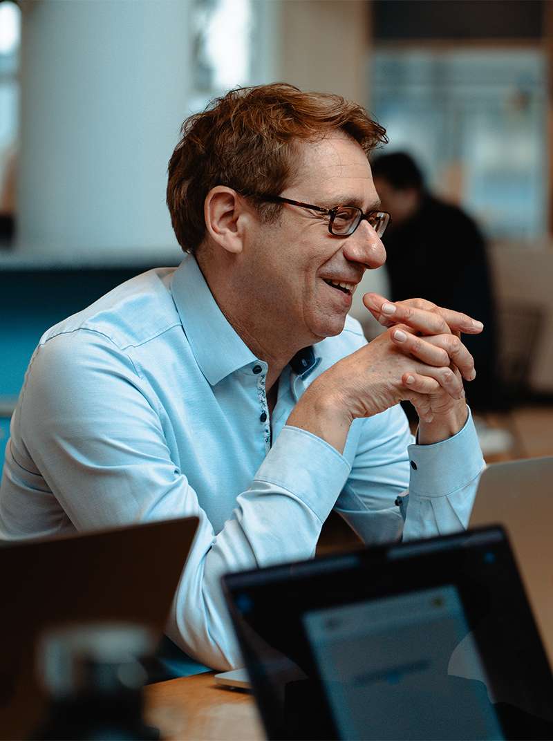 Lothar sitzt im Coworking-Space am Tisch und lacht während er die Hände locker gefaltet und die Ellenbogen auf dem Tisch aufgestützt hat.