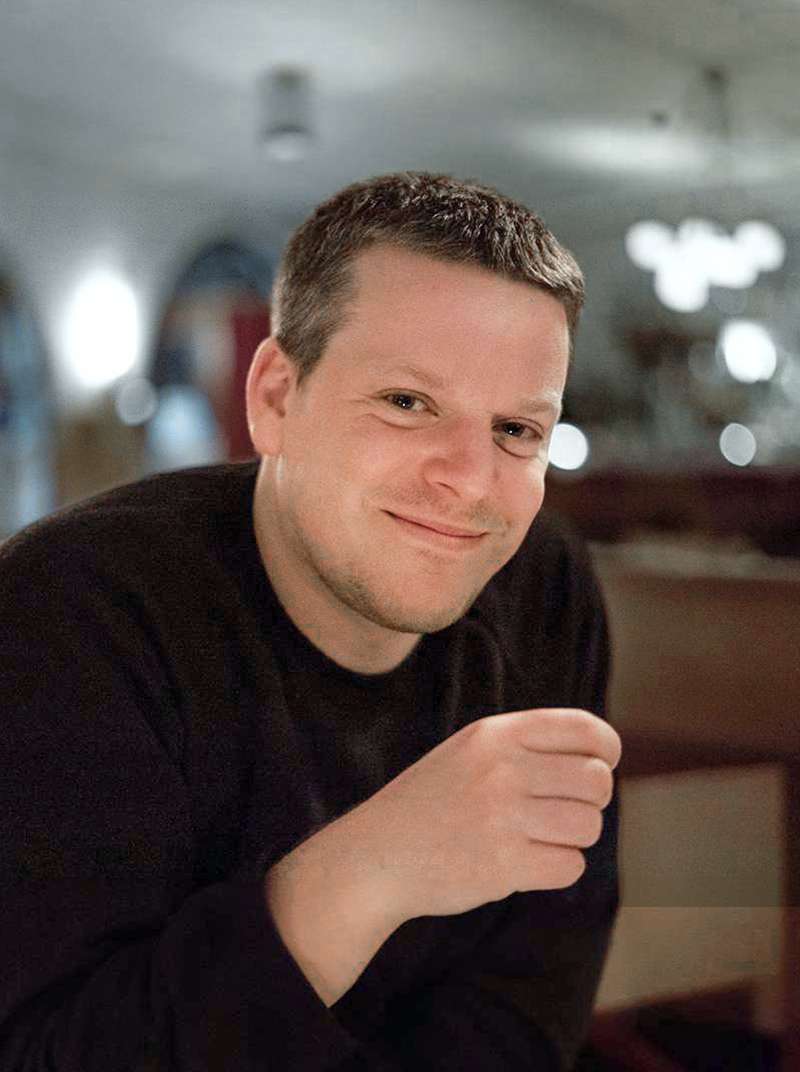 Markus sitzt lächelnd in einem Restaurant und hat die rechte Hand vor den Oberkörper gehoben.