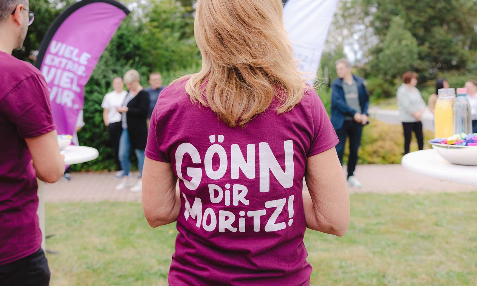 Rückenansicht einer Frau, die ein violettes T-Shirt mit der Aufschrift “Gönn dir Moritz” trägt.