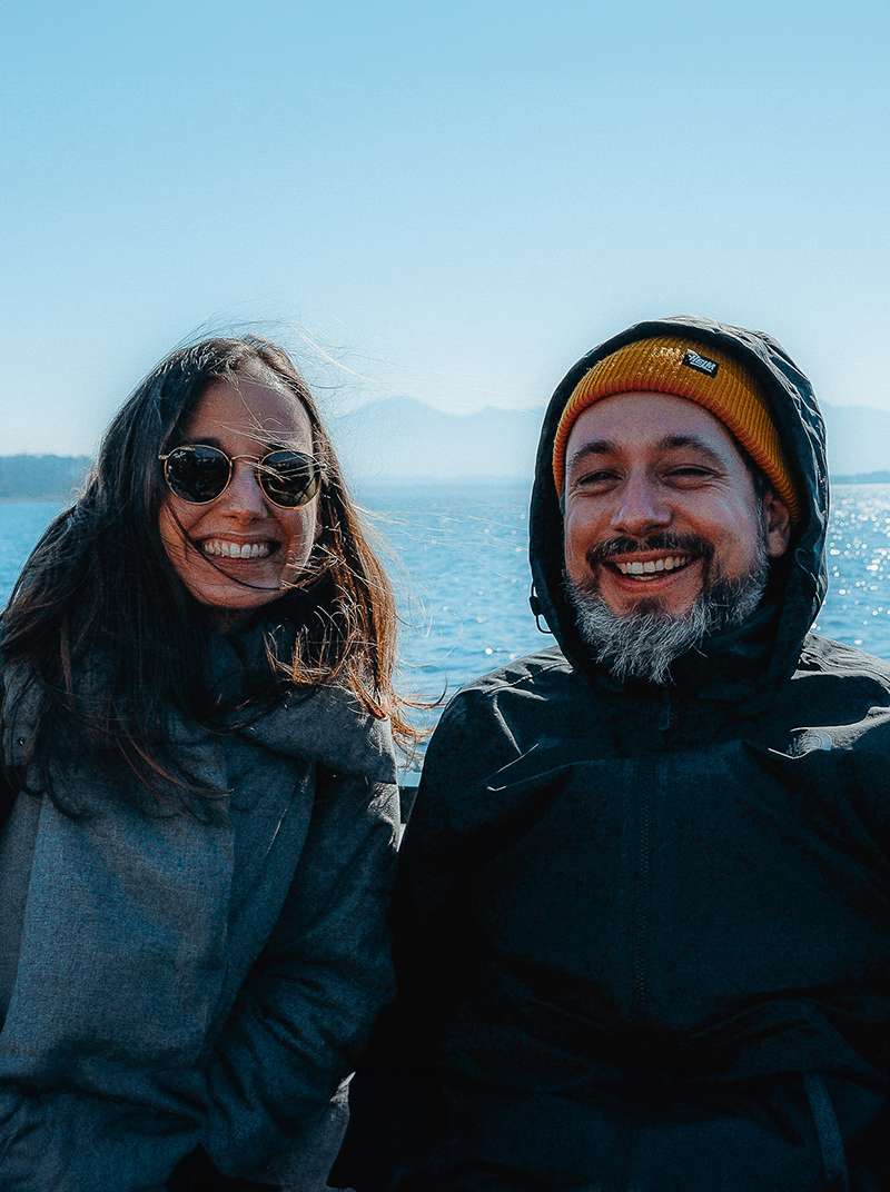 Sarah und Arne stehen in dicken Jacken an einem See und lachen in die Kamera. Die Sonne scheint.