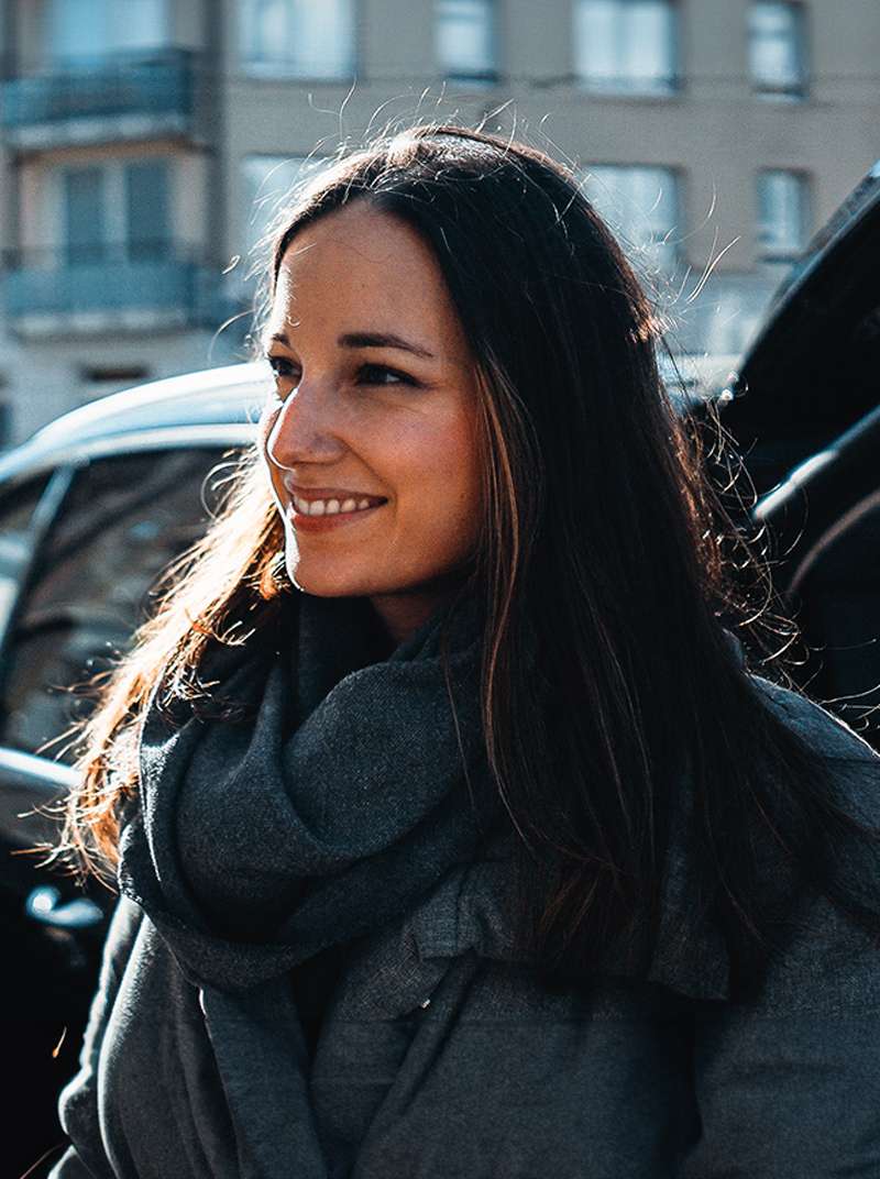 Sarah lächelt und ist von der Seite zu sehen. Sie trägt Winterjacke und Schal.