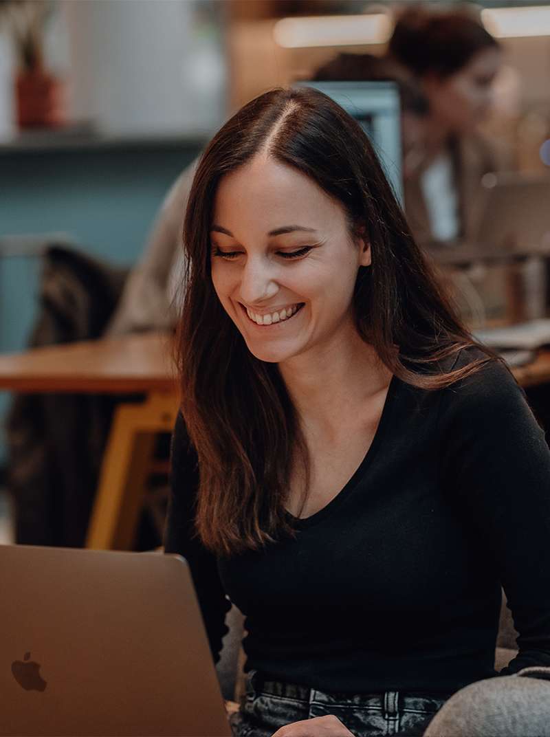 Sarah sitzt lachend hinter ihrem MacBook im Coworking-Space.