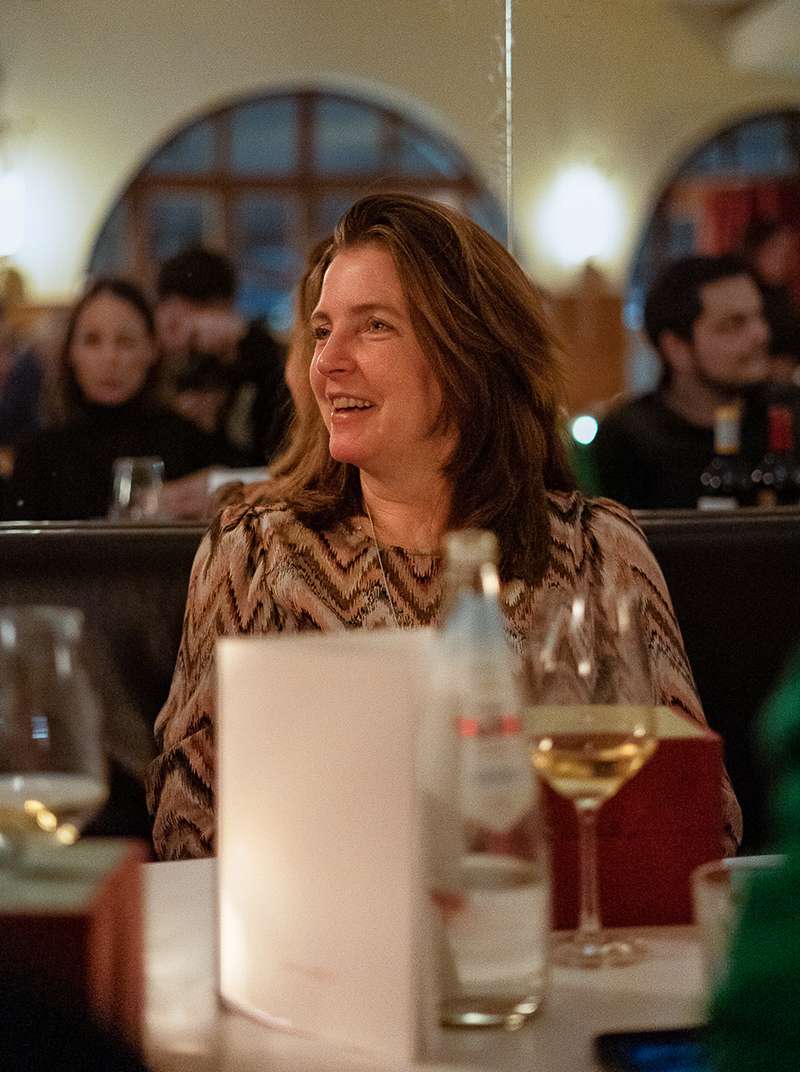 Siggi sitzt lachend an einem Tisch im Restaurant und hat ein Glas Weißwein vor sich stehen. Im Hintergrund sind leicht verschwommen Sarah und Amadeus zu sehen.